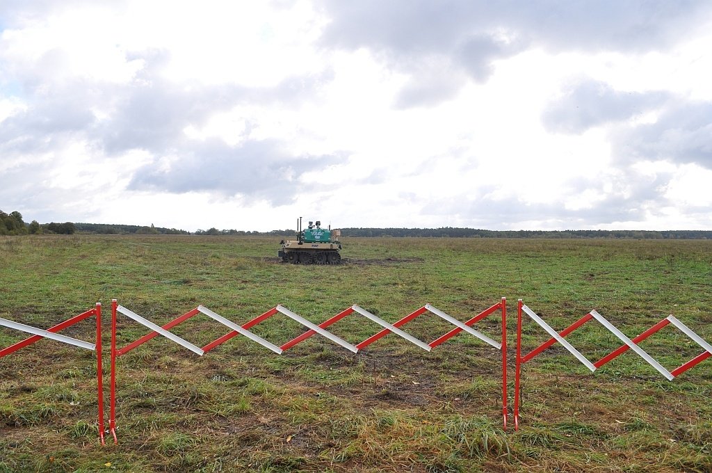 A marked line of red and white barriers running across open grassland, with a small vehicle in the far distance.