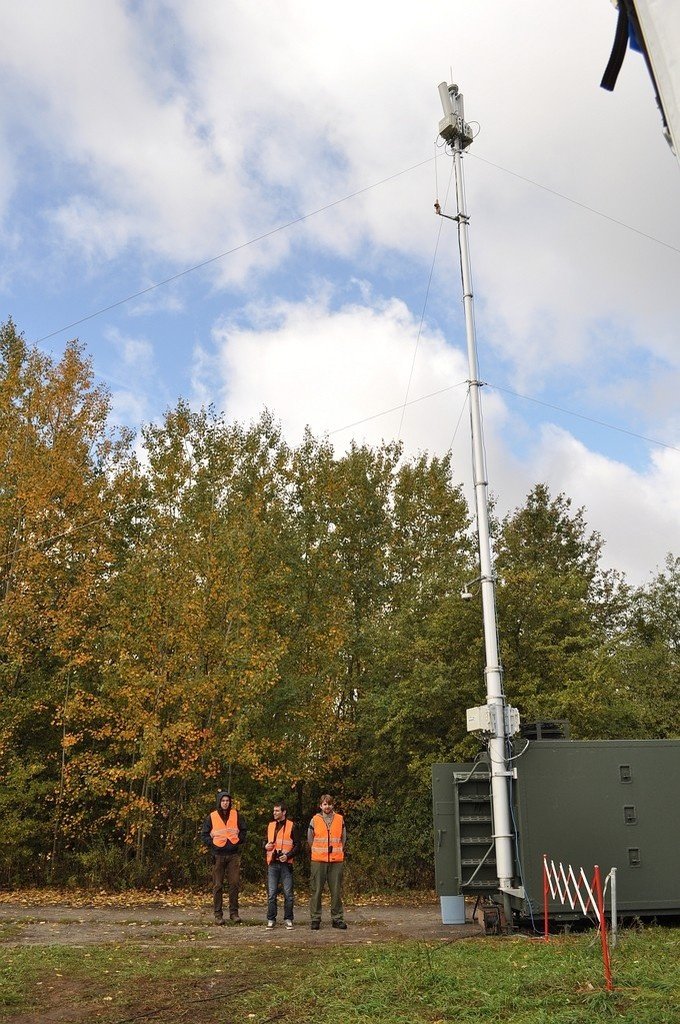 A slender lattice mast standing at the edge of a treeline, with a low building and a few figures at its base.