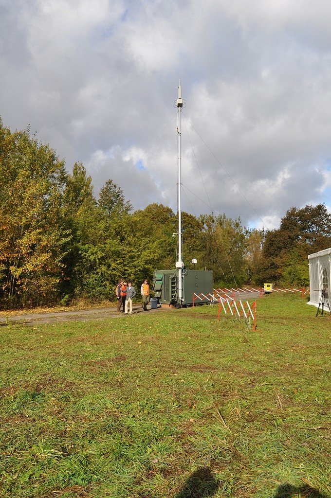 A portable lattice mast on a trailer standing at the edge of a mown field, guyed to the ground, with a small group of people beside it and lines of barriers behind.