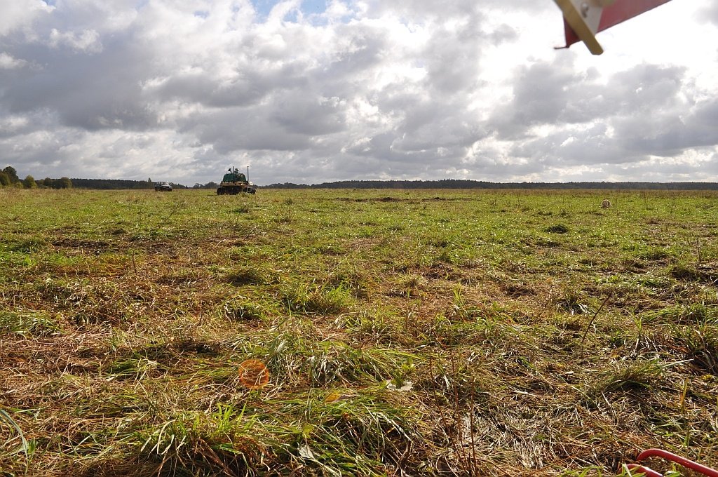 Wide mown grassland under broken cloud, a tracked platform working alone in the far distance and a barrier pole crossing the top of the frame.